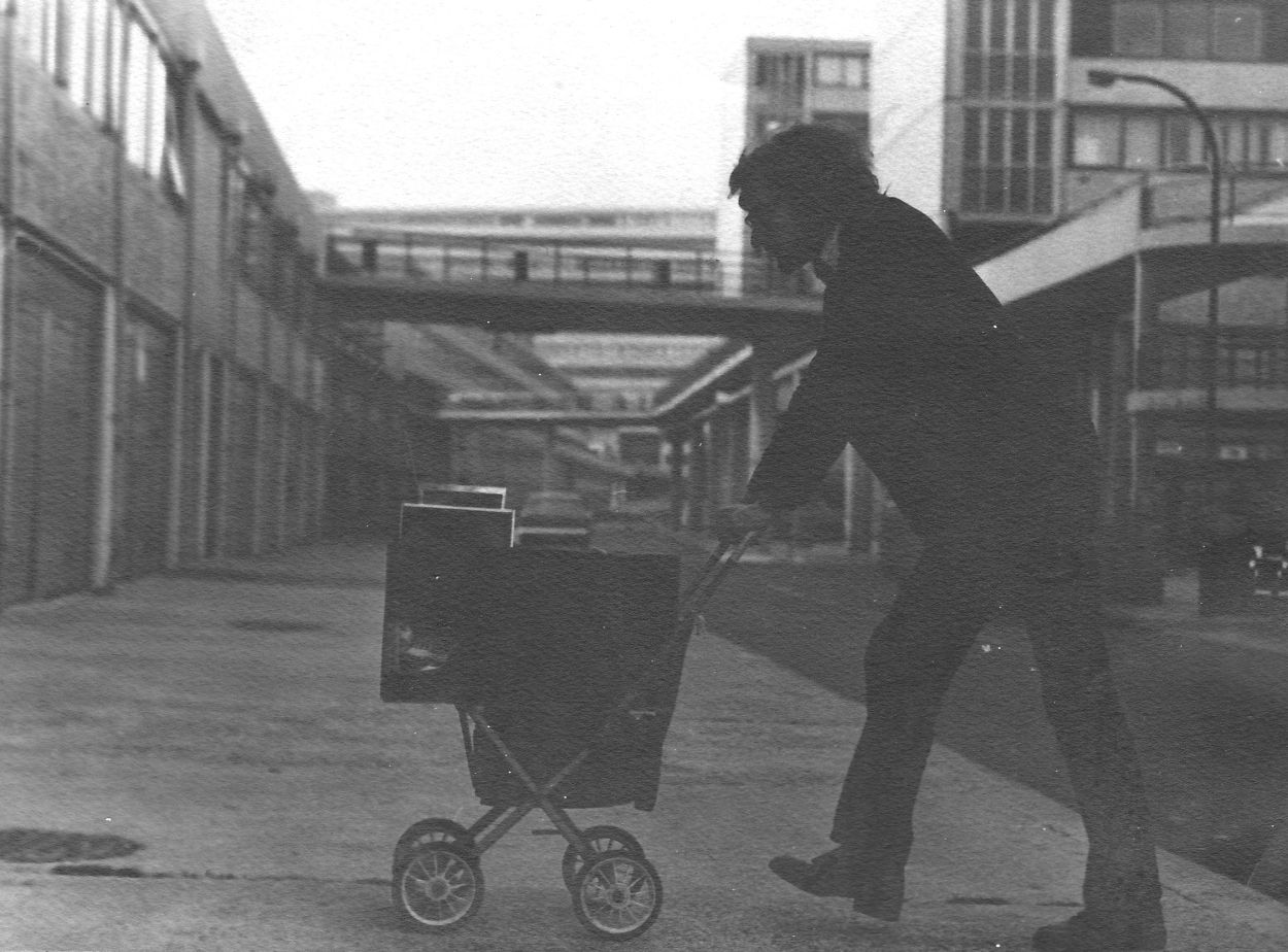Black and white image of man walking a pram through a housing estate..