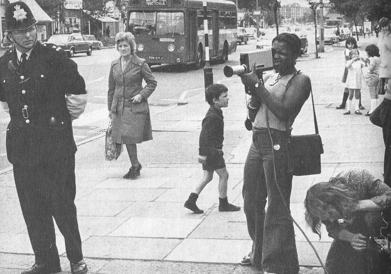 1980s black and white photo of a street scene with a young woman with a TV camera, a police officer, and a few pedestrians.