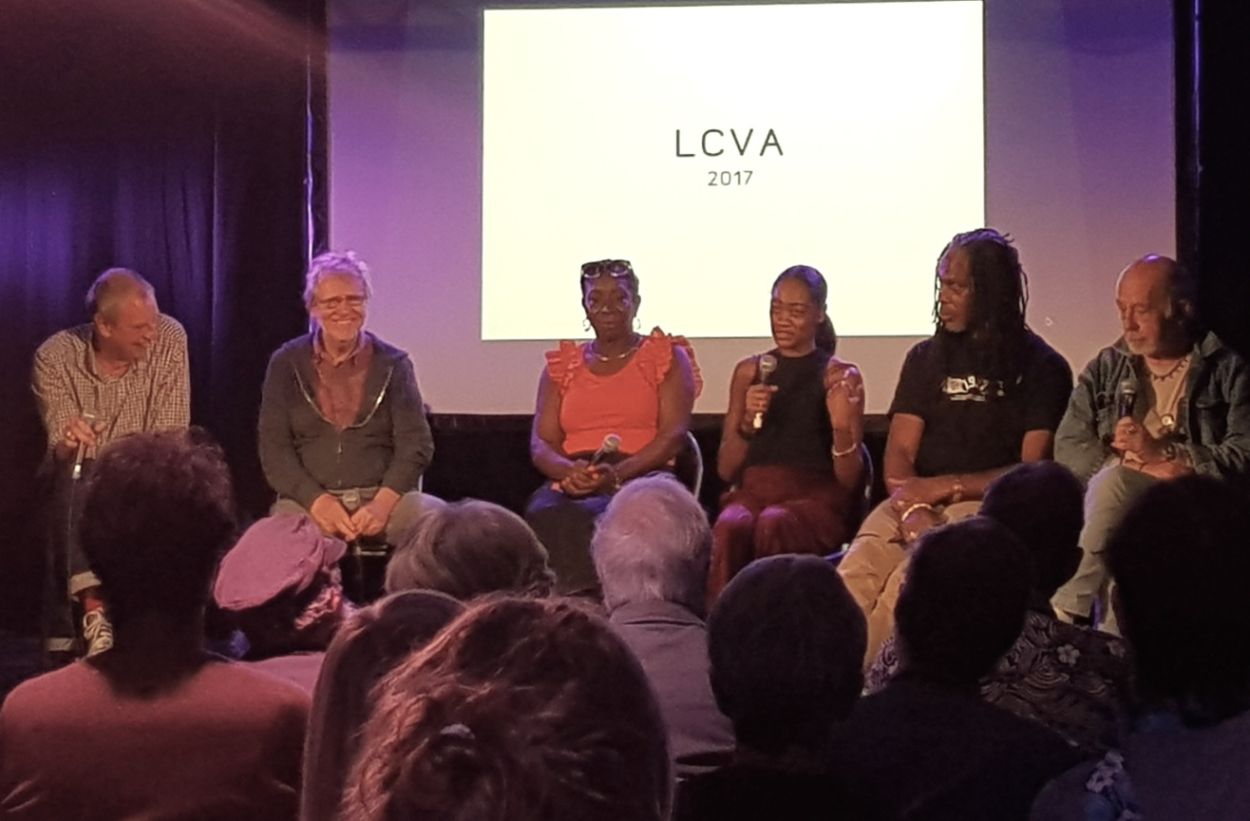Colour photo of a group of people sat talking infront of a crowd, with a screen behind them..